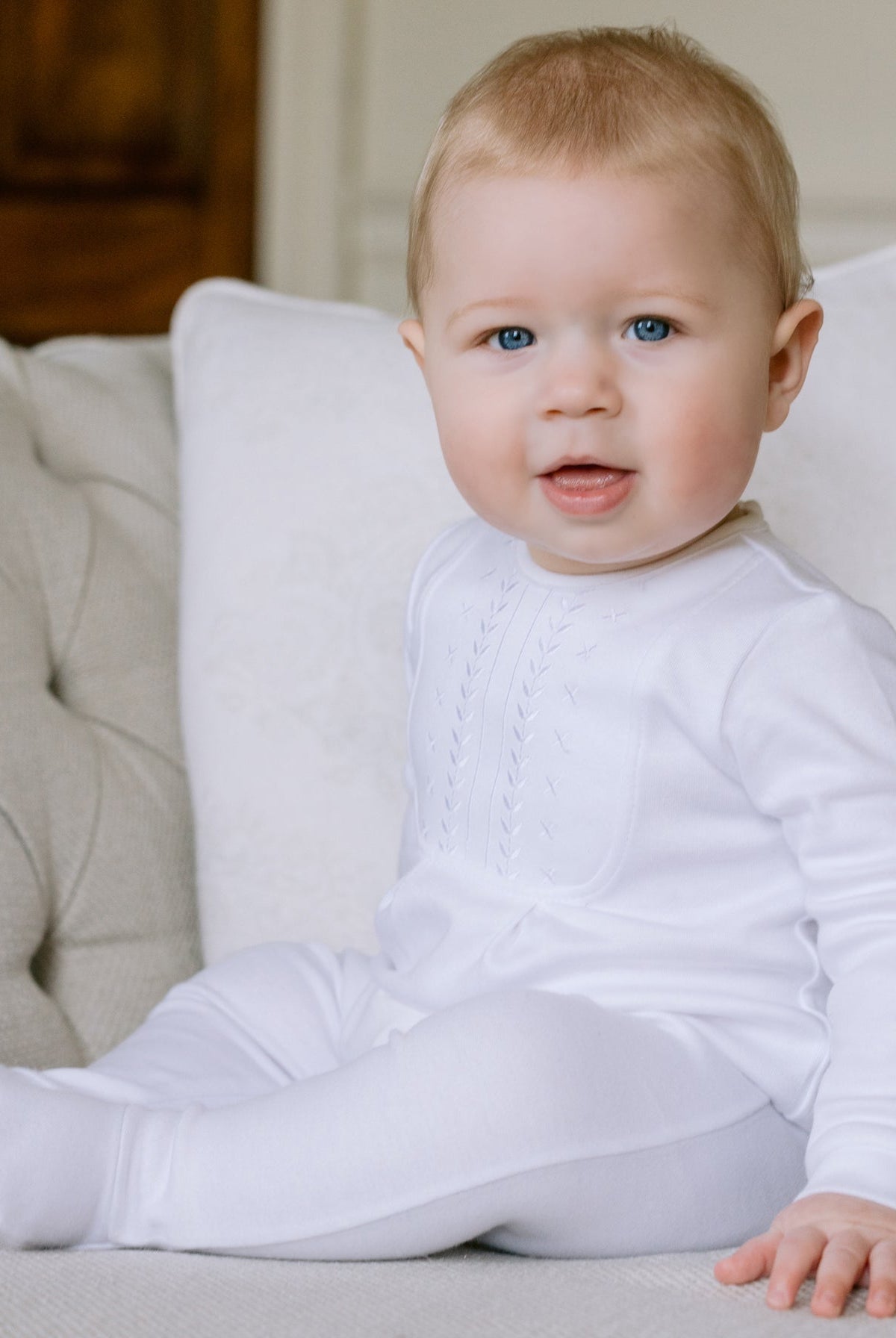 Baby in a white outfit sitting on a beige couch with a neutral background