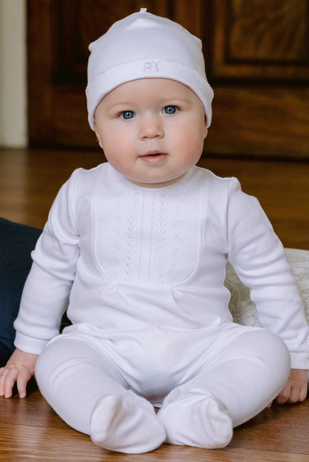 Baby in a white outfit sitting on a wooden floor