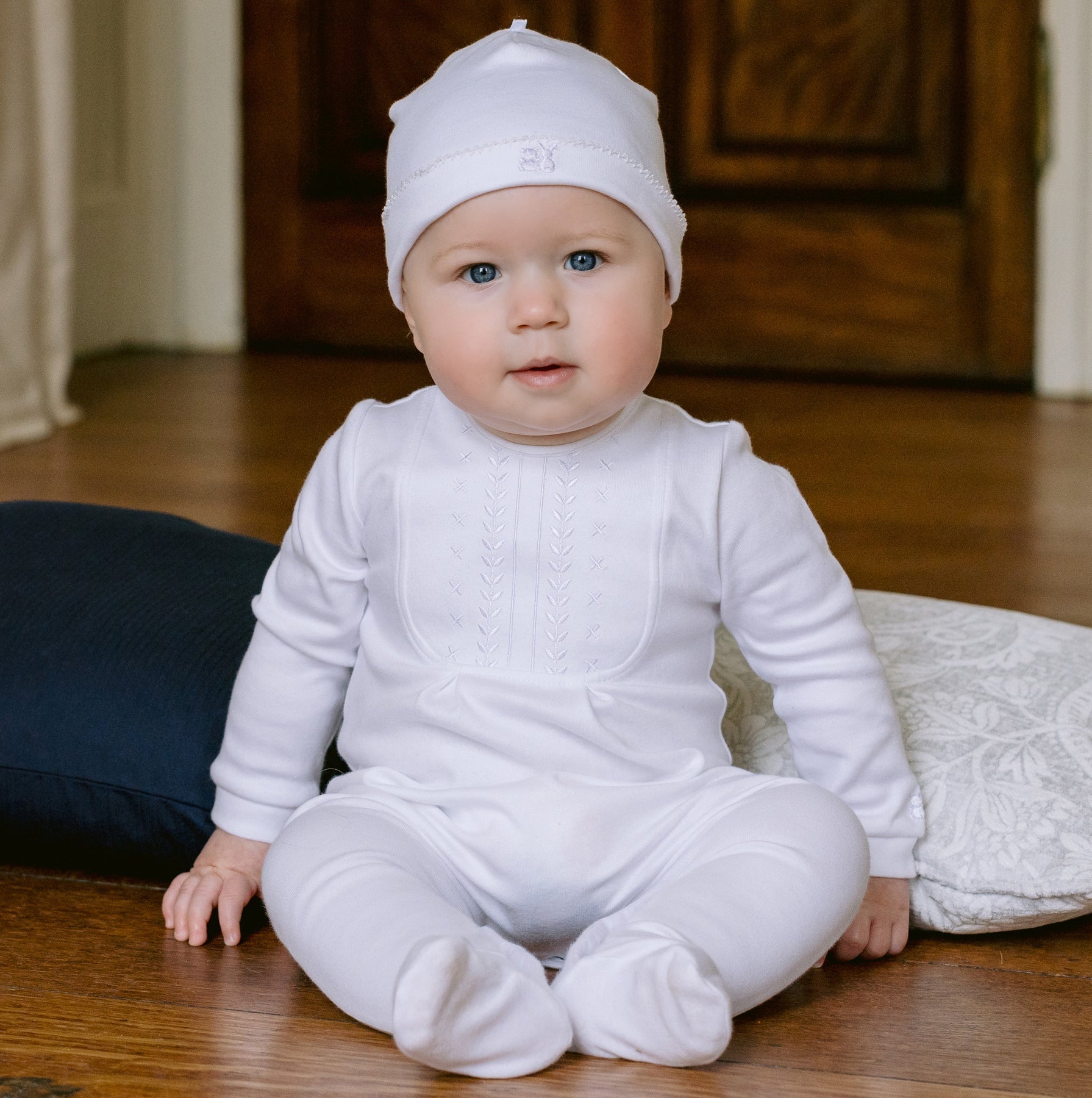 Baby in a white outfit sitting on a wooden floor