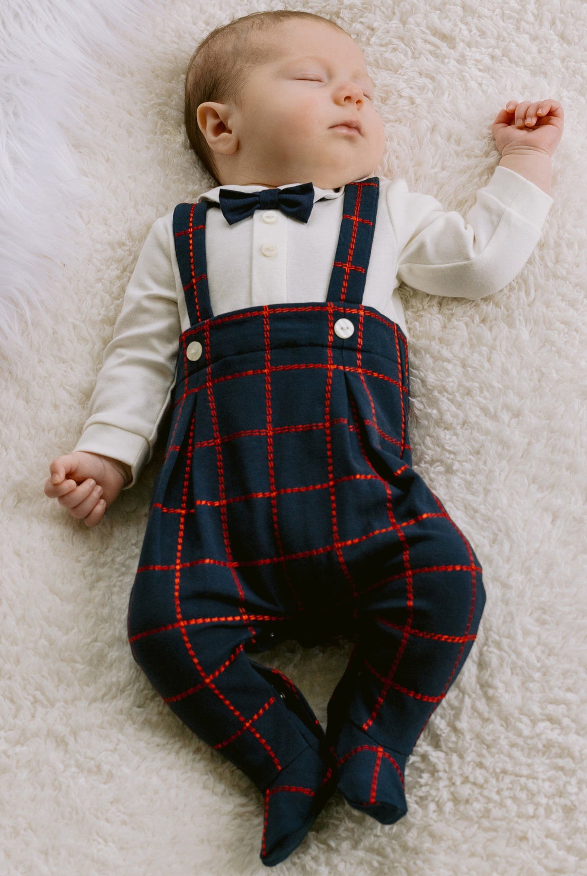 Newborn baby wearing a white shirt, black bow tie, and navy plaid overalls on a white fluffy blanket.