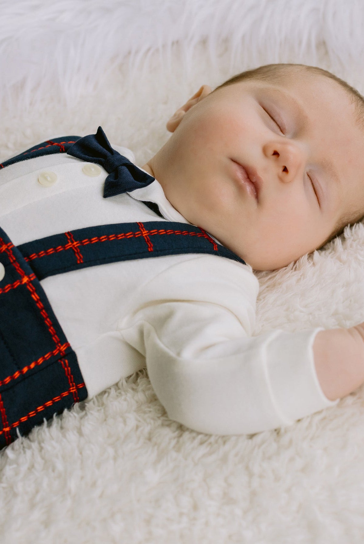 Newborn baby wearing a plaid outfit lying on a white fluffy surface