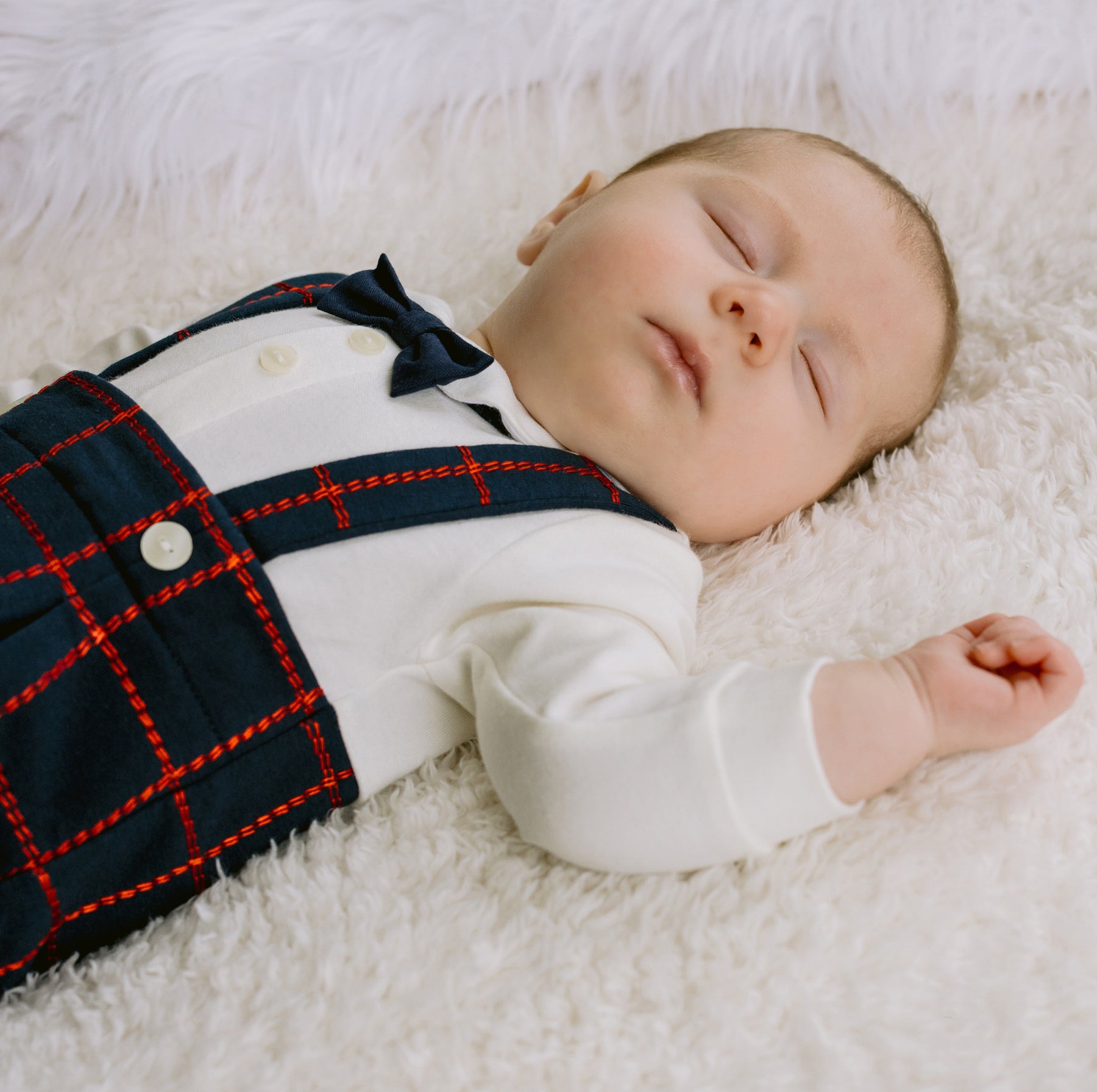 Newborn baby wearing a plaid outfit lying on a white fluffy surface