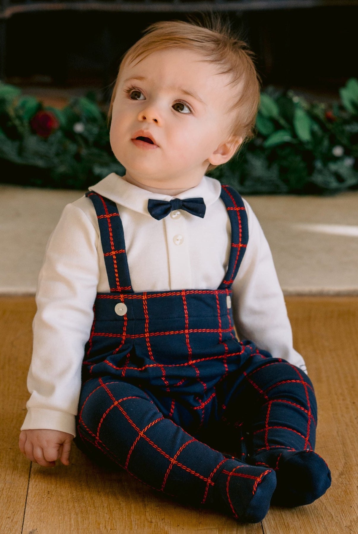Child wearing a navy plaid outfit with suspenders and a bow tie, sitting on a wooden floor.