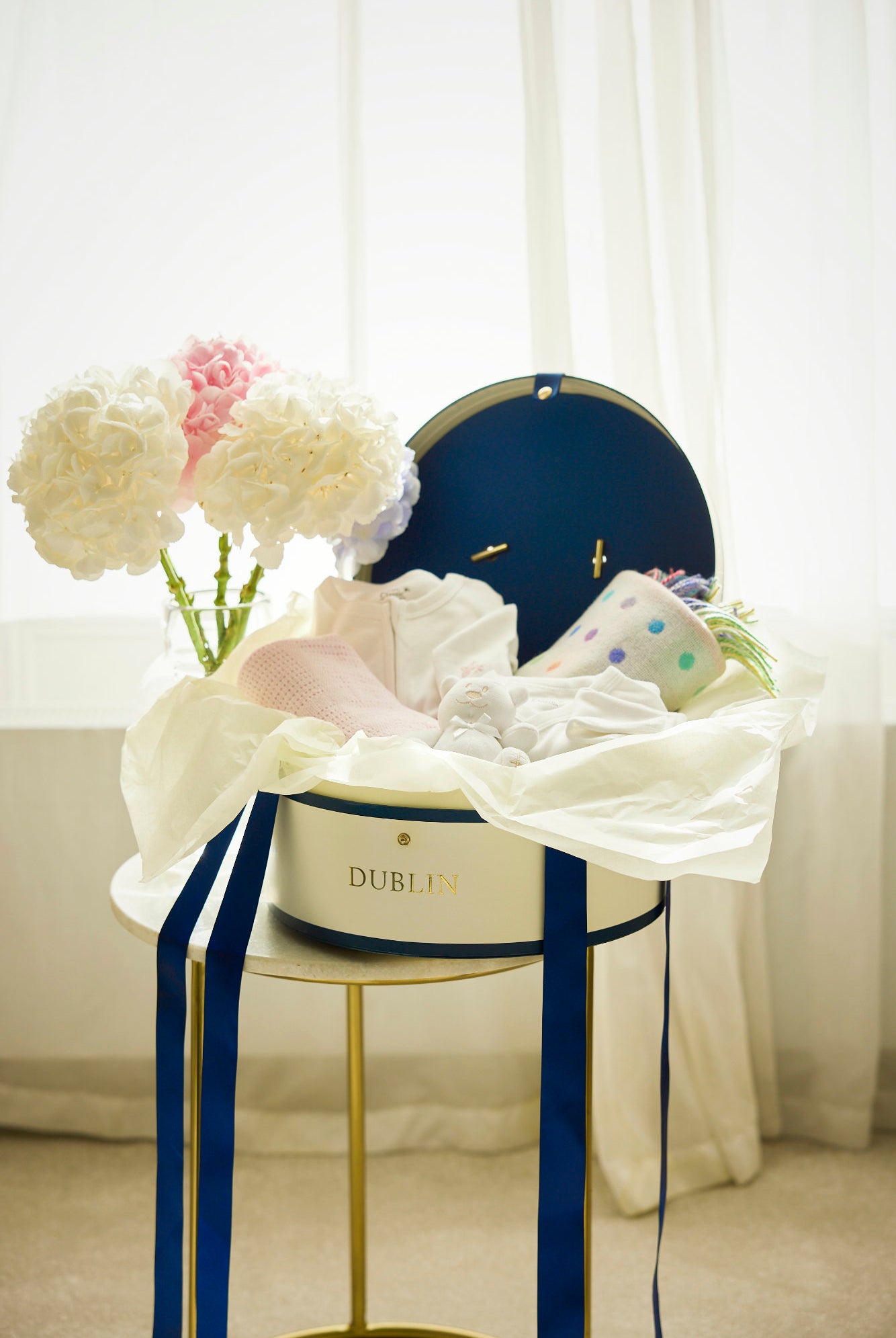 A baby gift set including a white and pink babygrow, a white and blue wrap over vest, a white and pink cellular baby shawl, a white lambswool blanket, a blue cuddly toy, and a classic hat box all displayed on a table with decorative ribbons and flowers.