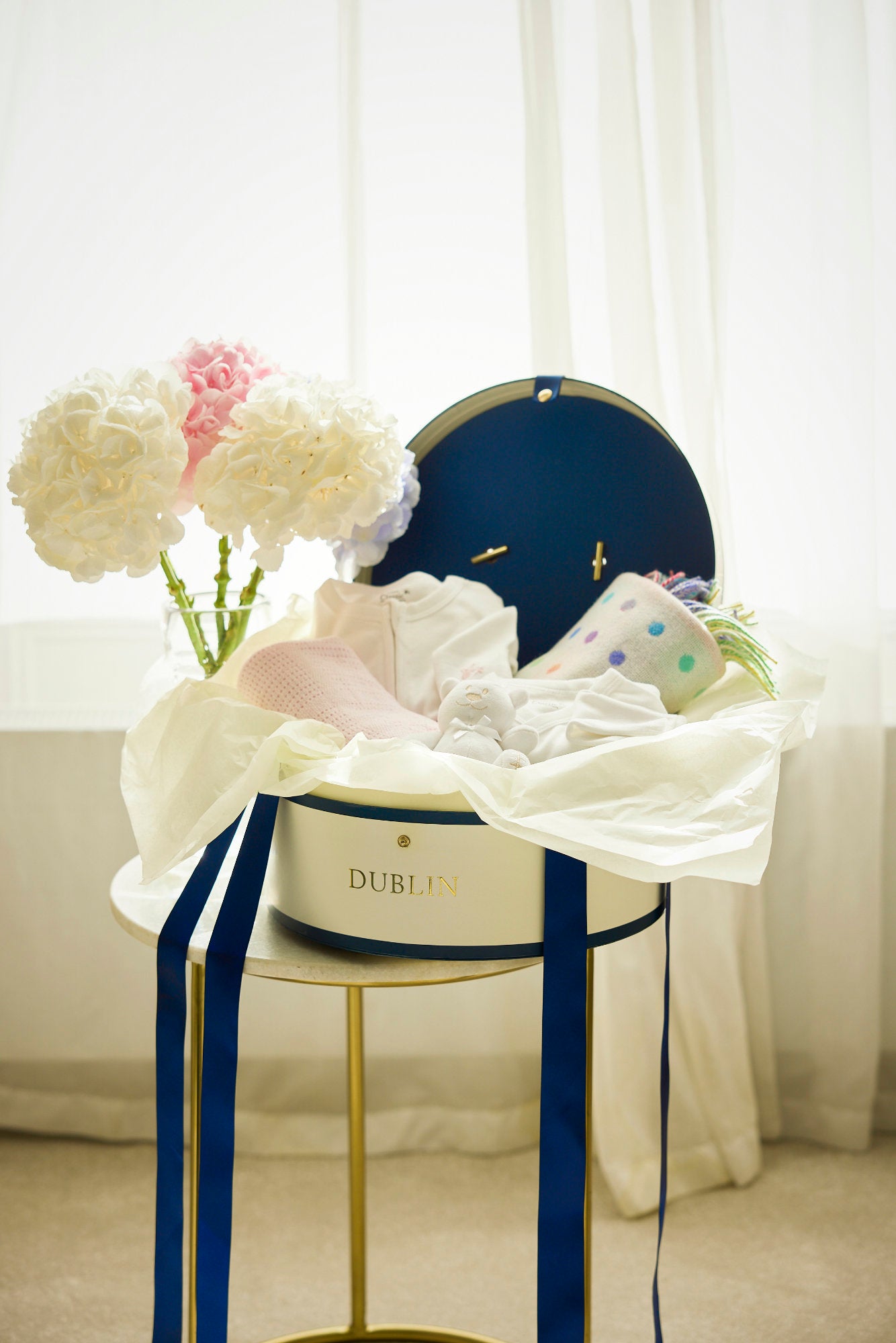 A baby gift set including a white and pink babygrow, a white and blue wrap over vest, a white and pink cellular baby shawl, a white lambswool blanket, a blue cuddly toy, and a classic hat box all displayed on a table with decorative ribbons and flowers.