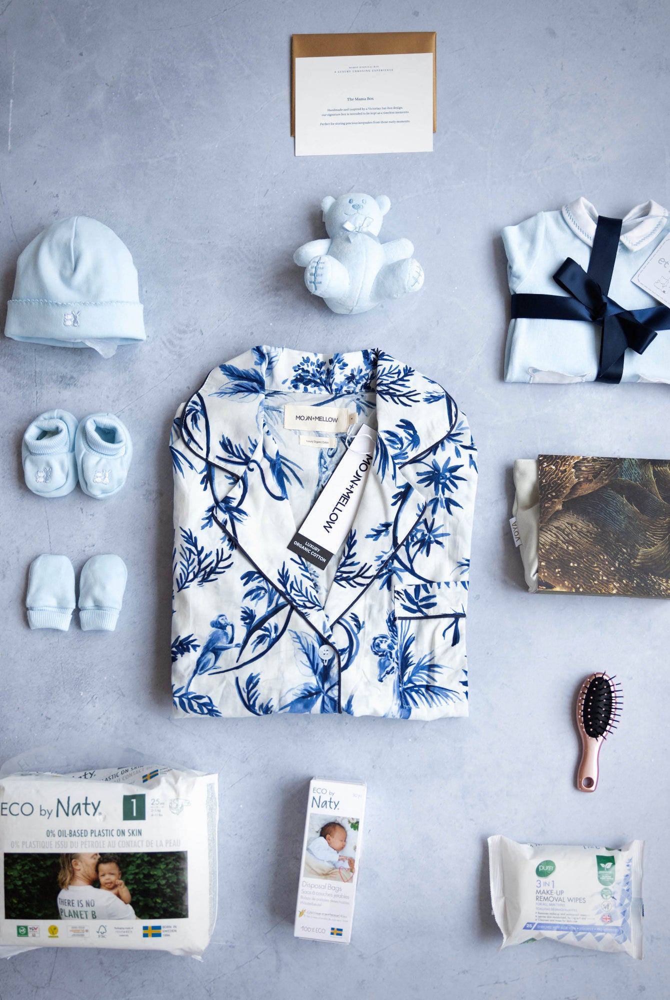 A set of baby and maternity related items in blue, including a nightdress, baby clothes, a matching hat, a cuddly toy, a mini hair brush, nappies, wipes, and a keepsake box.