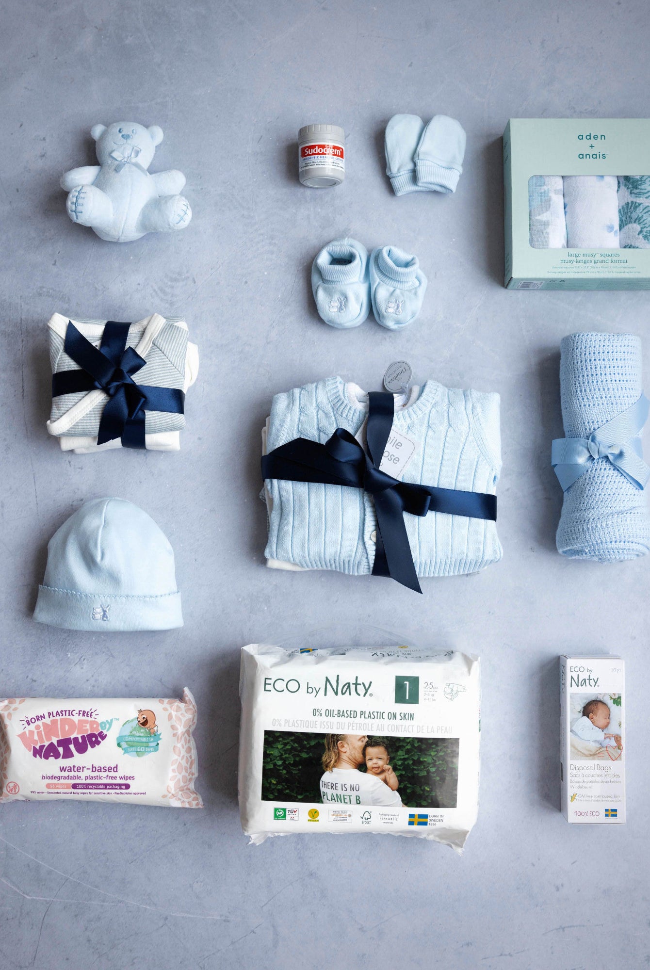 A collection of baby items laid out on a grey background, including a baby hat, sleepwear, a cardigan, long sleeve vests, a hat with a bow, a cellular blanket, a cuddly toy, muslins, a pack of nappies, and a pack of wipes, all in a branded packaging.