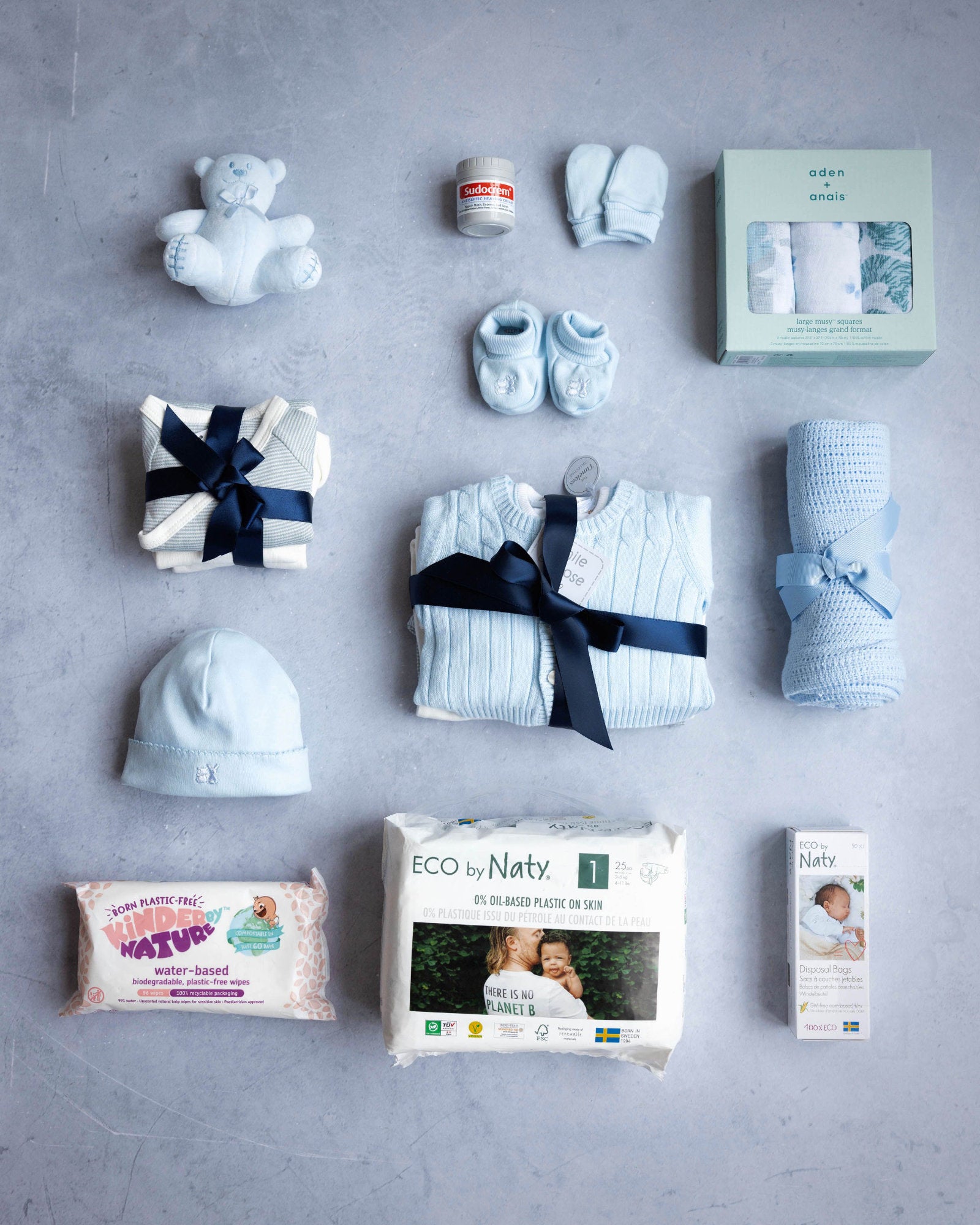 A collection of baby items laid out on a grey background, including a baby hat, sleepwear, a cardigan, long sleeve vests, a hat with a bow, a cellular blanket, a cuddly toy, muslins, a pack of nappies, and a pack of wipes, all in a branded packaging.