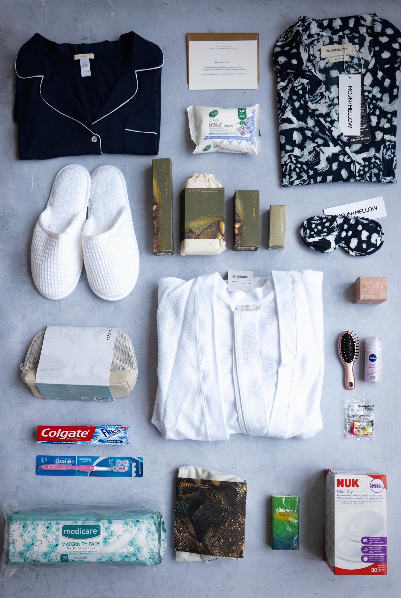 A collection of baby and maternity items including a nightdress, sleepshirts, pyjama set, eye cream, facecloths, facial set, shower gel, shampoo and conditioner, body lotion, mama oil, hand cream, and a candle, displayed neatly on a grey background.