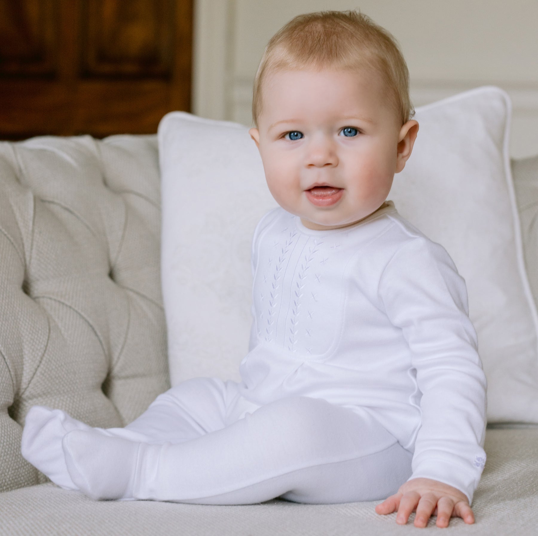 Baby in a white outfit sitting on a beige couch with a neutral background