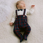 Newborn baby wearing a white shirt, black bow tie, and navy plaid overalls on a white fluffy blanket.