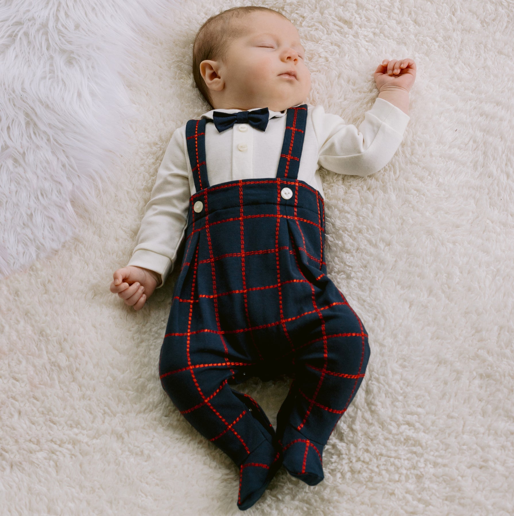 Newborn baby wearing a white shirt, black bow tie, and navy plaid overalls on a white fluffy blanket.