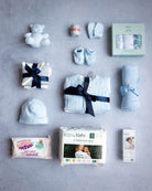 A collection of baby items laid out on a grey background, including a baby hat, sleepwear, a cardigan, long sleeve vests, a hat with a bow, a cellular blanket, a cuddly toy, muslins, a pack of nappies, and a pack of wipes, all in a branded packaging.