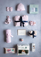 A baby gift set displayed with various items including a pink beanie, three pink sleepsuits, a pink cardigan, long sleeve vests, a hat, scratch mitts, booties, a cellular blanket, a cuddly toy, muslins, a pack of nappies, friendly wipes, and disposable nappy bags, all neatly arranged on a grey background.