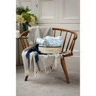 A baby gift set displayed on a wooden rocking chair including a gray babygro, blue hat, and a beige lambswool blanket with fringes, placed in a cozy room setting.
